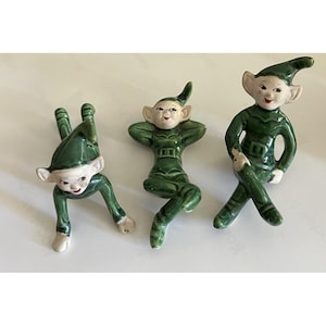 May include: Three ceramic elf figurines with glossy green outfits and pointed hats. One elf is positioned in a crawling pose, one is lying on its back with hands behind its head, and one is sitting upright with legs crossed, all featuring painted faces.
