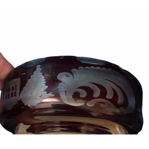 May include: A ruby red glass bowl with etched white designs. The bowl features a winter scene with a house and a tree, along with a decorative floral pattern. The rim of the bowl has a scalloped edge.