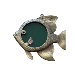 May include: A gold-colored metal fish picture frame with a green glass insert. The fish has a detailed design and a hanging loop on the top.