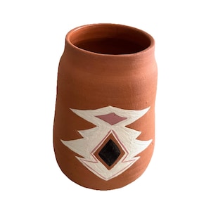 May include: A terracotta-colored ceramic vase with a unique, hand-painted design. The design features a central black diamond shape, framed by pink and white geometric patterns. The vase has a cylindrical shape with a slightly wider base.