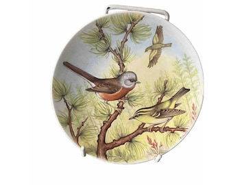 May include: A white ceramic plate with a hand-painted design of three birds perched on a branch of a pine tree. The birds are yellow, brown, and gray with red accents. The plate has a white metal stand.