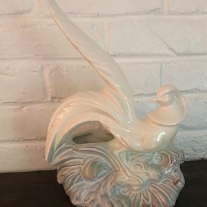May include: A white ceramic bird figurine with a long tail, perched on a leafy base. The bird has a delicate, stylized design and a light iridescent glaze.