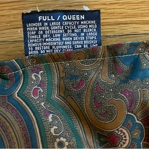 Op de afbeelding: Een donkerblauw stoffen waslabel met witte tekst die luidt FULL / QUEEN, LAUNDER IN LARGE CAPACITY MACHINE, WARM WASH, GENTLE CYCLE, USING MILD SOAP OR DETERGENT, DO NOT BLEACH, TUMBLE DRY, LOW SETTING, bevestigd aan een textiel met een blauwgroen paisley-patroon.
