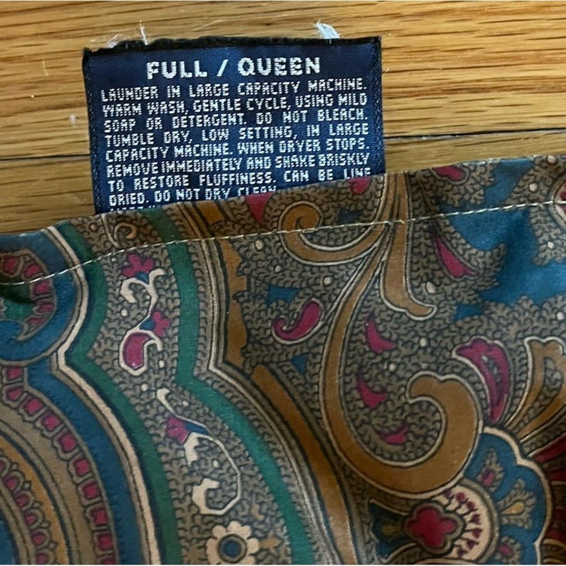 Op de afbeelding: Een donkerblauw stoffen waslabel met witte tekst die luidt FULL / QUEEN, LAUNDER IN LARGE CAPACITY MACHINE, WARM WASH, GENTLE CYCLE, USING MILD SOAP OR DETERGENT, DO NOT BLEACH, TUMBLE DRY, LOW SETTING, bevestigd aan een textiel met een blauwgroen paisley-patroon.