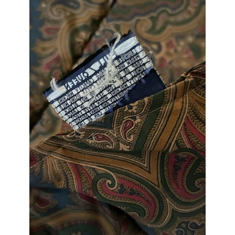 Op de afbeelding: Een kledingstuk van stof met een complex paisley-patroon in tinten donkerblauw, olijfgroen en dieprood. Een rechthoekig kledinglabel is aan de stof bevestigd, met witte tekst op een donkerblauwe achtergrond met gerafelde randen.