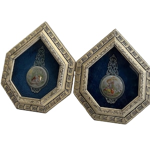 May include: Two pentagonal wall frames with ornate gold-toned metal borders and deep blue velvet backings. Each frame contains a central circular porcelain medallion featuring a colorful painted scene of figures in a landscape with intricate metal filigree.