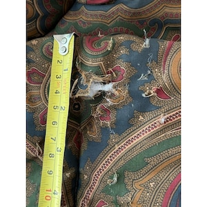 Op de afbeelding: Een stof met een paisley-patroon in donkerblauw, groen en rood met een zichtbare scheur in het materiaal. Een geel meetlint is verticaal tegen de stof geplaatst en toont een afmeting van ongeveer 10 cm in lengte bij de beschadiging.
