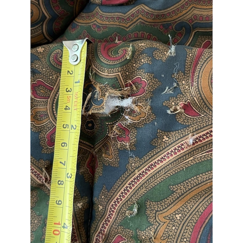 Op de afbeelding: Een stof met een paisley-patroon in donkerblauw, groen en rood met een zichtbare scheur in het materiaal. Een geel meetlint is verticaal tegen de stof geplaatst en toont een afmeting van ongeveer 10 cm in lengte bij de beschadiging.