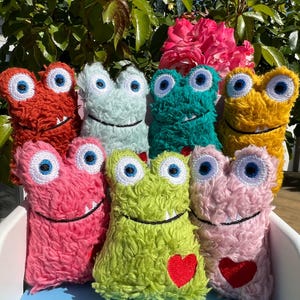 Könnte beinhalten: Eine Sammlung von sieben Plüschmonster-Spielzeugen aus zotteligem, strukturiertem Stoff in verschiedenen Farben, darunter Rot, Hellblau, Türkis, Gelb, Pink, Limettengrün und Hellrosa. Jedes Monster hat zwei große weiße Augen mit blauen Pupillen.