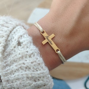 May include: A gold cross charm bracelet with the name "Faustine" engraved on it. The bracelet is made of a beige cord and is worn on a person's wrist.