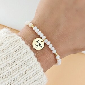 Pode incluir: Uma pulseira de contas com pequenas contas brancas facetadas e detalhes em tom dourado. Um pingente redondo de metal dourado pende do centro, gravado com o nome Jeanne em letra cursiva e um pequeno gráfico de duas pegadas de bebé.