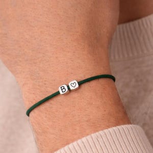 Peut inclure: Un bracelet vert foncé avec deux petits cubes argentés. L'un porte la lettre "B", l'autre un cœur. Le bracelet est porté au poignet.