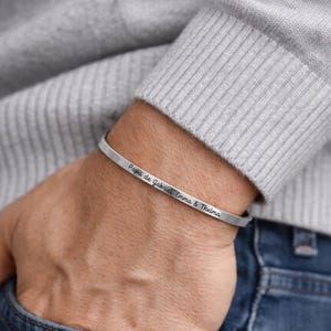 May include: A silver-colored cuff bracelet with the inscription "Papa de Gabriel Emma & Thelma" is worn on a wrist. The person is wearing a gray sweater and blue jeans. The bracelet is a simple, elegant design.