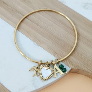 May include: Gold bangle bracelet with charms, including a heart with a white center, a small bird, a letter 'A', and two green beads.
