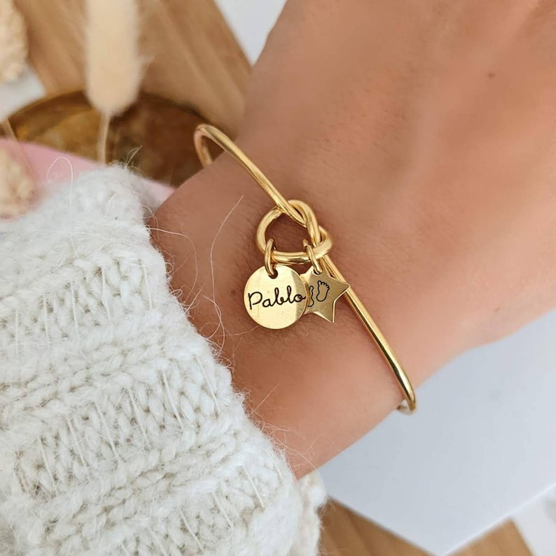 May include: Gold bangle bracelet with a knot detail and two charms. One charm is a round disc with the name "Pablo" engraved on it. The other charm is a star with a footprint design.