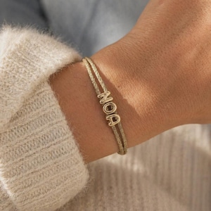 Op de afbeelding: Een goudkleurige koordarmband met drie metalen letterkralen die de naam NOA spellen. De armband bestaat uit twee dunne, getextureerde gouden koorden die de letters op hun plaats houden, wat zorgt voor een eenvoudig en strak ontwerp om de pols.