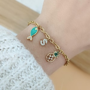 Peut inclure: Bracelet en chaîne dorée avec des breloques, dont un poisson vert, un ananas et une lettre 'S' avec des strass.