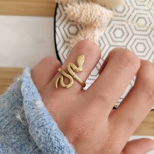 Könnte beinhalten: Ein goldfarbener Schlangenring. Der Ring umschlingt den Finger, wobei sich Kopf und Schwanz der Schlange treffen. Der Ring hat eine strukturierte Oberfläche und wird an einem Finger getragen. Der Hintergrund umfasst einen blauen Ärmel, eine gemusterte Oberfläche und Trockenblumen.