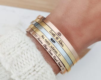 May include: A stack of four thin metal cuff bracelets in rose gold, gold, silver, and copper tones. Each bracelet is engraved with custom text including names like Scott, Alba, Maïssa, Aliya, Tarès, and Riyad, alongside small icons.