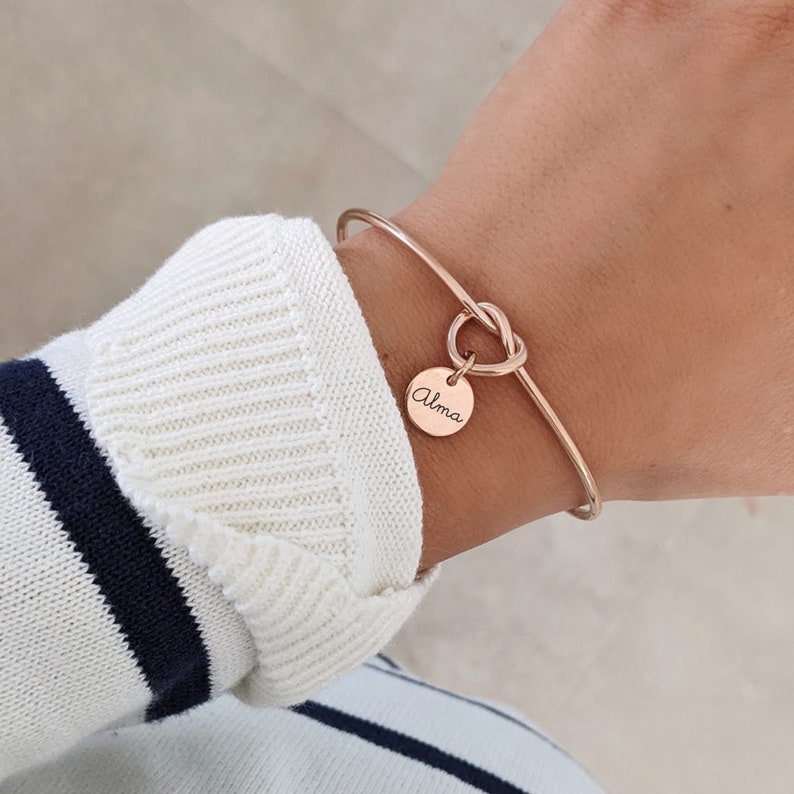 May include: A rose gold bangle bracelet with a knot detail and a round charm that says "Alma".