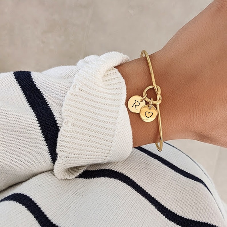 May include: Gold bangle bracelet with a knot detail and two charms. One charm is a round disc with the letter "R" engraved on it. The other charm is a heart with a small heart engraved inside it.