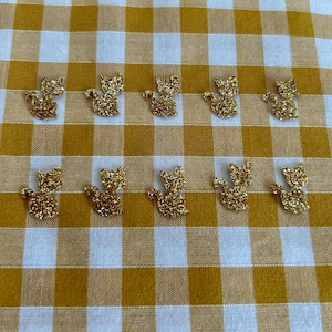 Peut inclure: Dix embellissements dorés en forme de chat en paillettes, disposés sur un tissu à carreaux jaune et blanc. Les figurines de chat sont détaillées avec des particules de paillettes visibles, créant un effet scintillant.
