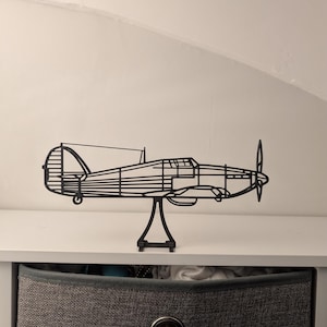 Op de afbeelding: Een zwarte metalen vliegtuigsculptuur met een propeller en een standaard. Het decoratieve stuk wordt tentoongesteld op een witte plank, met een grijze opbergbak eronder. Het vliegtuig is een gedetailleerde, gestileerde weergave.