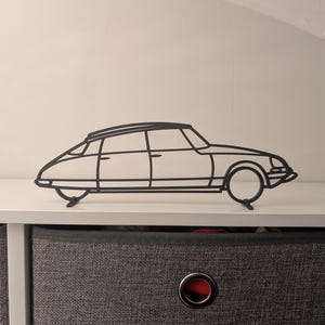 Può includere: Una silhouette di un'auto classica in metallo nero, probabilmente una Citroën DS, montata su uno scaffale bianco sopra un cassetto in tessuto grigio con un occhiello rosso.