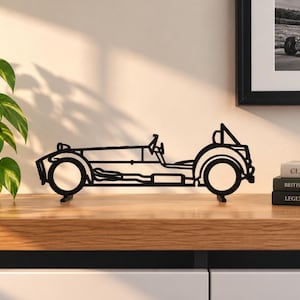 May include: A black metal wire-frame sculpture of a vintage open-top sports car. The silhouette features a long bonnet, exposed wheels, a steering wheel, and a roll bar. The design is flat and stands on two small base supports on a wooden surface.