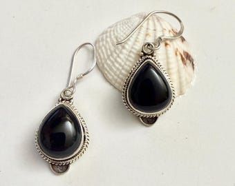 May include: A pair of silver earrings with teardrop-shaped, black gemstones. The gemstones are set in a silver frame with a rope-like border. The earrings are displayed on a white surface next to a seashell.