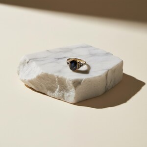 K&ouml;nnte beinhalten: Ein goldener Ring mit einem gro&szlig;en ovalen, dunkelblauen Edelstein und kleineren Akzentsteinen. Der Ring wird auf einem wei&szlig;en Marmorblock vor neutralem Hintergrund pr&auml;sentiert, um sein Vintage-Design und seine eleganten Details hervorzuheben.