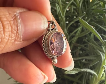 May include: A silver pendant with an oval, light pink gemstone. The pendant has a decorative border and a small, round bead at the bottom. The background is a blurred view of green foliage.