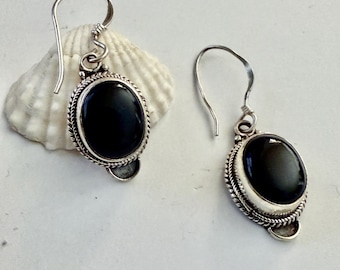 May include: A pair of silver earrings with oval, black gemstones. The gemstones are set within a silver frame with a twisted rope design. The earrings have a simple hook closure. The earrings are displayed on a white surface.