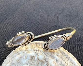 May include: A silver-toned cuff bracelet with two oval-shaped, light-colored gemstones. The gemstones are set in ornate bezels with a twisted rope design. The bracelet is displayed on a seashell against a dark background.