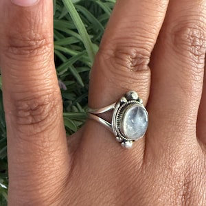 Könnte beinhalten: Ein silberner Ring mit einem ovalen, irisierenden Edelstein wird auf einer Hand gezeigt. Der Ring hat eine detaillierte, verzierte Fassung mit kleinen dekorativen Elementen an den Seiten. Der Hintergrund zeigt grünes Laub.