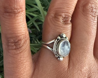 May include: A silver ring with an oval, iridescent gemstone is displayed on a hand. The ring has a detailed, ornate setting with small decorative elements on the sides. The background features green foliage.