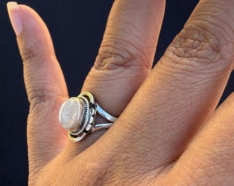 May include: A silver ring featuring a large, oval, pale stone. The stone is set in a decorative bezel with a floral design. The ring's band is plain, polished silver. The ring is on a person's finger.