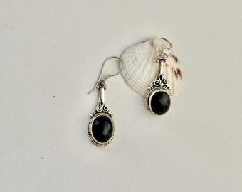 May include: A pair of silver dangle earrings with oval, black gemstones. The earrings have a decorative design at the top and a simple hook closure. The earrings are displayed on a white surface with a seashell.