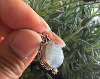 May include: A silver pendant with an oval, iridescent moonstone. The pendant has a detailed, beaded border and a small, round accent at the bottom. The background features green foliage.