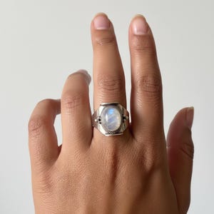 Könnte beinhalten: Ein silberner Ring mit einem ovalen, schillernden Mondstein. Der Ring hat ein rechteckiges Band mit dekorativen Details an den Seiten. Der Mondstein ist in einer Zargenfassung gefasst. Der Ring befindet sich an einem Finger.
