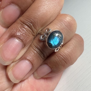 Könnte beinhalten: Ein silberner Ring mit einem ovalen, irisierenden blauen Edelstein. Der Ring hat ein dekoratives, wirbelndes Design an den Seiten des Edelsteins. Der Ring befindet sich an einem Finger.