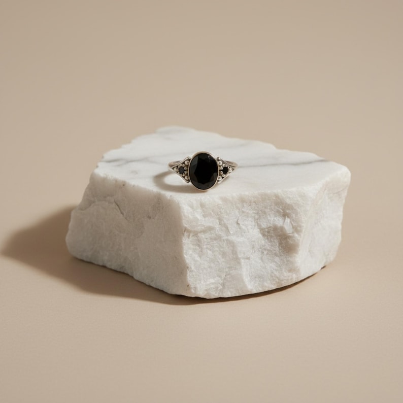 K&ouml;nnte beinhalten: Ein silberner Ring mit einem gro&szlig;en ovalen schwarzen Stein in der Mitte, flankiert von kleineren schwarzen Steinen. Der Ring liegt auf einer wei&szlig;en Marmoroberfl&auml;che vor einem neutralen Hintergrund. Das Design des Rings ist aufwendig und detailliert.