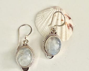 May include: A pair of silver drop earrings with oval, iridescent moonstone cabochons. The stones are set in a detailed, rope-like border. The earrings are displayed on a white surface with a seashell in the background.