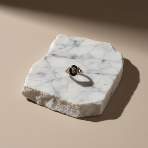K&ouml;nnte beinhalten: Ein goldener Ring mit einem ovalen, dunklen Edelstein in der Mitte und kleineren Steinen an den Seiten. Der Ring liegt auf einer wei&szlig;en Marmorplatte vor einem neutralen Hintergrund. Das Design des Rings weist aufwendige Details auf.