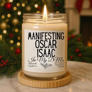 May include: A lit candle in a glass jar with a label that reads "MANIFESTING OSCAR ISAAC In My DM's". The candle sits on a wooden coaster. The background features a Christmas tree with lights and ornaments.