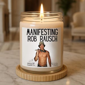 Może przedstawiać: Płonąca świeca z wosku sojowego w przezroczystym szklanym słoiku z białą etykietą z napisem "MANIFESTING ROB RAUSCH" oraz zdjęciem osoby z wężem. Świeca stoi na okrągłej, żebrowanej drewnianej podstawce.