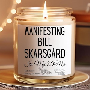 May include: A white candle with a black label that reads "MANIFESTING BILL SKARSGÅRD In My DM's". The label also includes the text "SisterSister Holiday Boutique" and "100% Soy Wax Burning Time 50-60 Hrs".