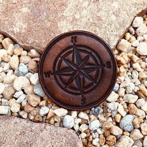 Peut inclure: Un dessous de verre rond en cuir marron avec un motif de rose des vents. La rose des vents présente les points cardinaux : Nord, Sud, Est et Ouest, avec une étoile au centre. Le dessous de verre est posé sur un lit de petits cailloux multicolores.