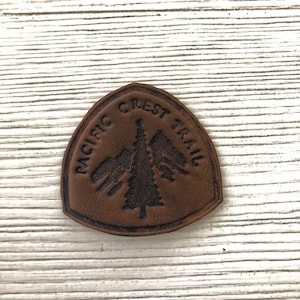 May include: A brown leather patch shaped like a shield with a debossed design featuring a central pine tree, mountain silhouettes on either side, and the text PACIFIC CREST TRAIL arched across the top portion of the textured surface.