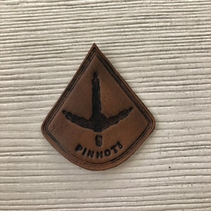 May include: A brown leather patch with a black silhouette of a bird's foot and the text "PINHOTT" in black.
